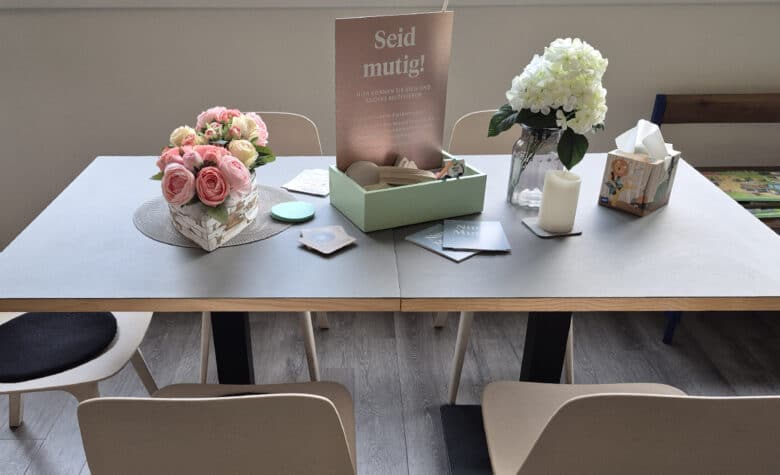 Ein Tisch mit mehreren Gegenständen: Blumen, Zettel, einer Kerze. Auf einem Schild steht: Seid mutig!
